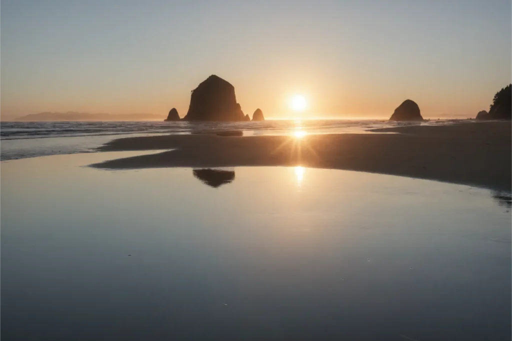 The sun setting over Cannon Beach, casting a warm glow and creating reflections on the wet sand, ideal for evening strolls and playful activities with Corgis.